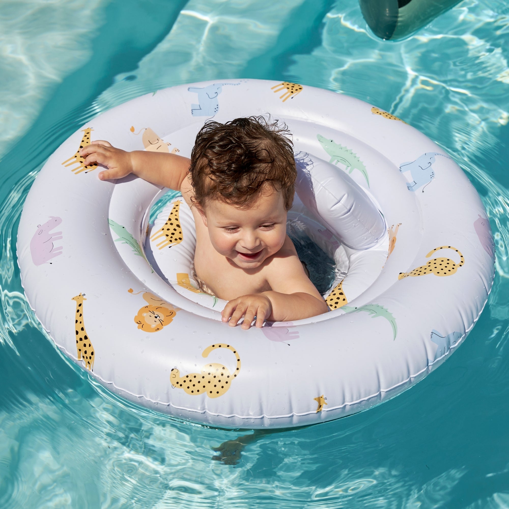 Happy child who is looking away in a clear blue pool in a white baby float with animal print on it.