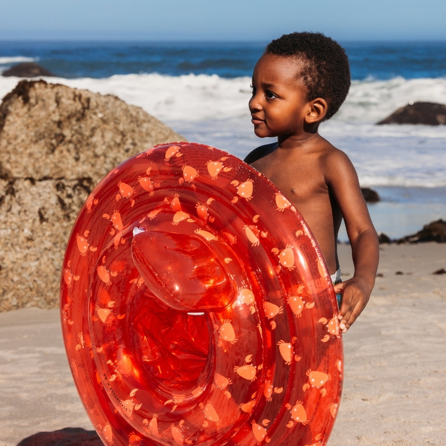 Child holding a transparant red baby float with crabs on it on a beach with ocean waves in the background