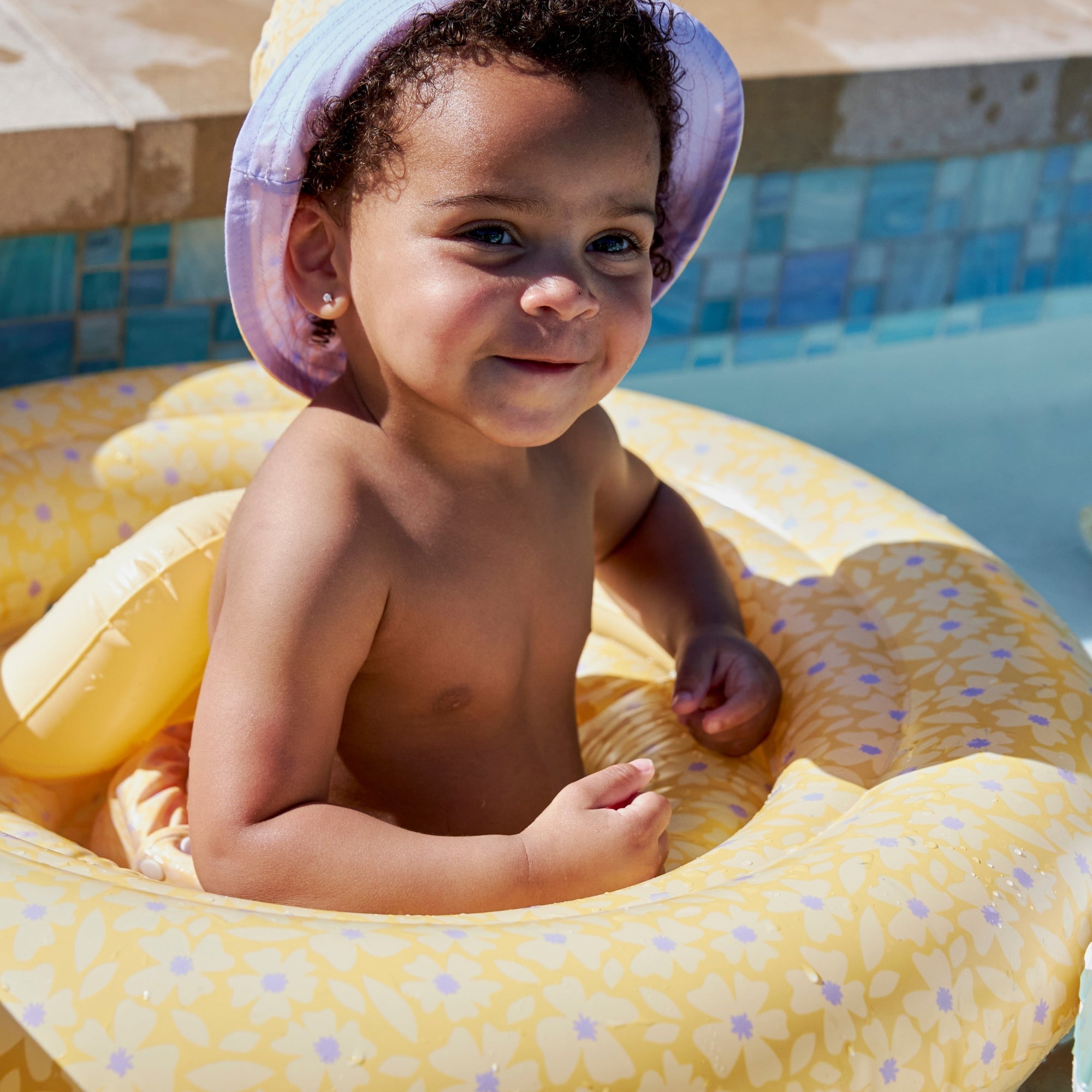 Small child in a pool in a yellow baby float with flowers and purple polka dots on it and purple hat