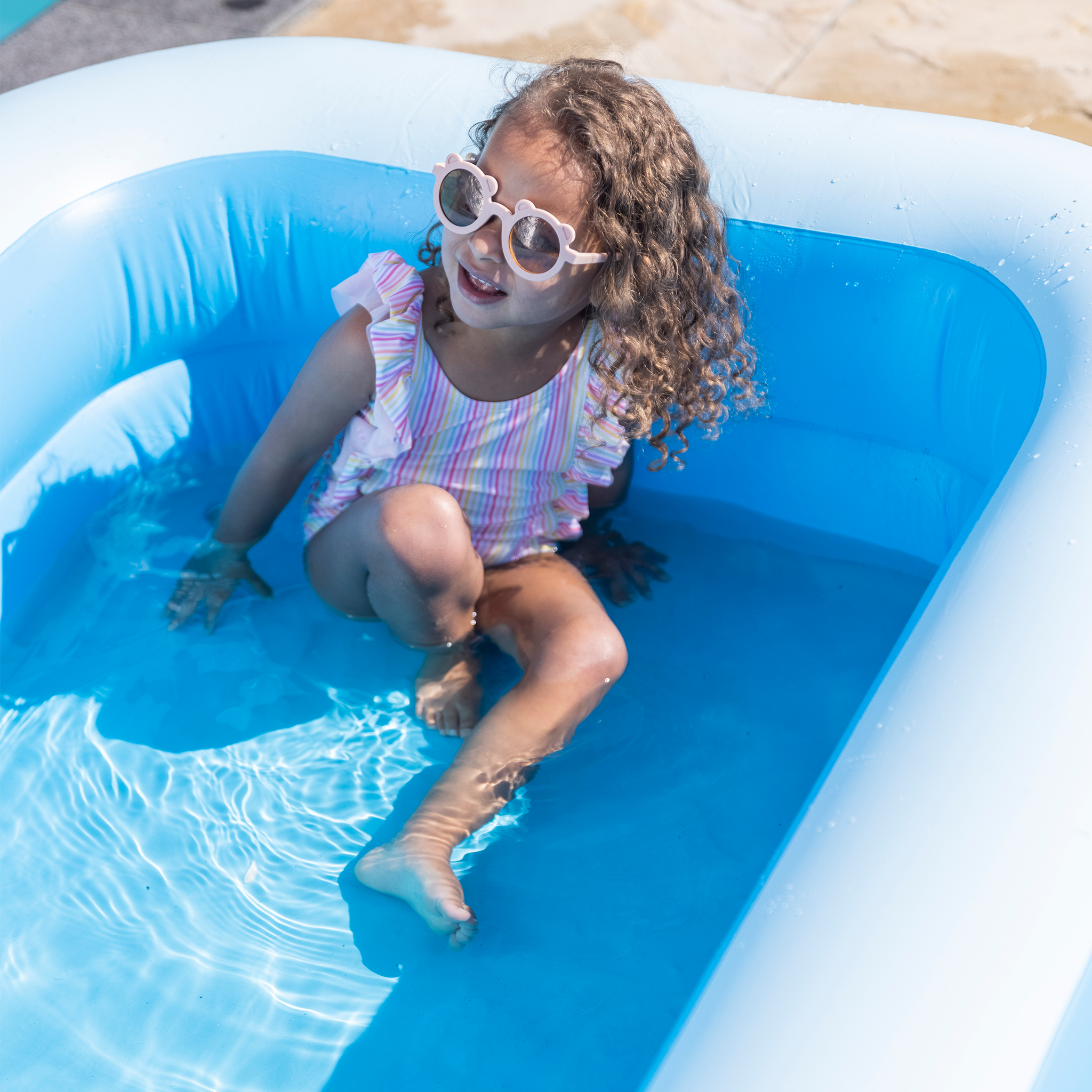 Girl in a colorful swimsuit and sunglasses sitting in a blue inflatable pool.