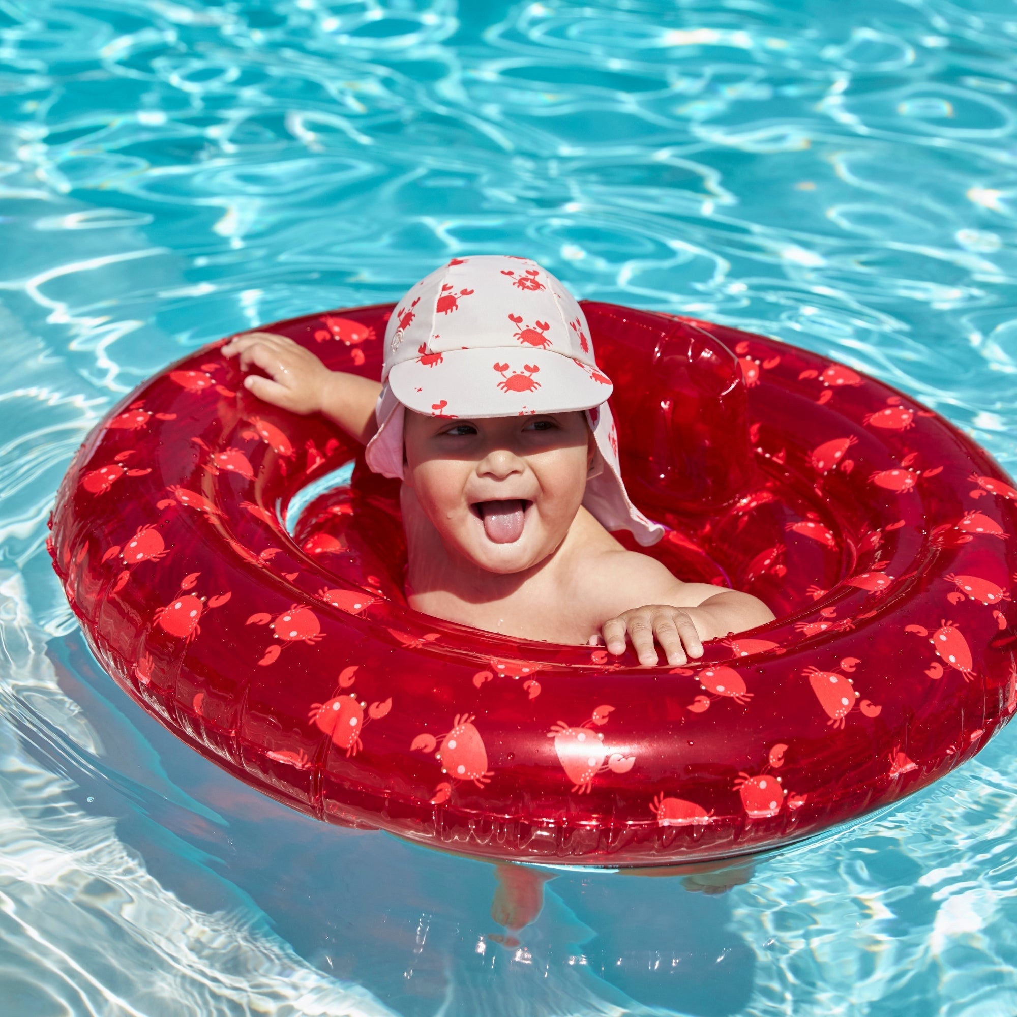 Happy child with his tongue out his mouth wearing a beige cap with crabs on it in a transparant red baby float with crabs on it in a pool with clear water.
