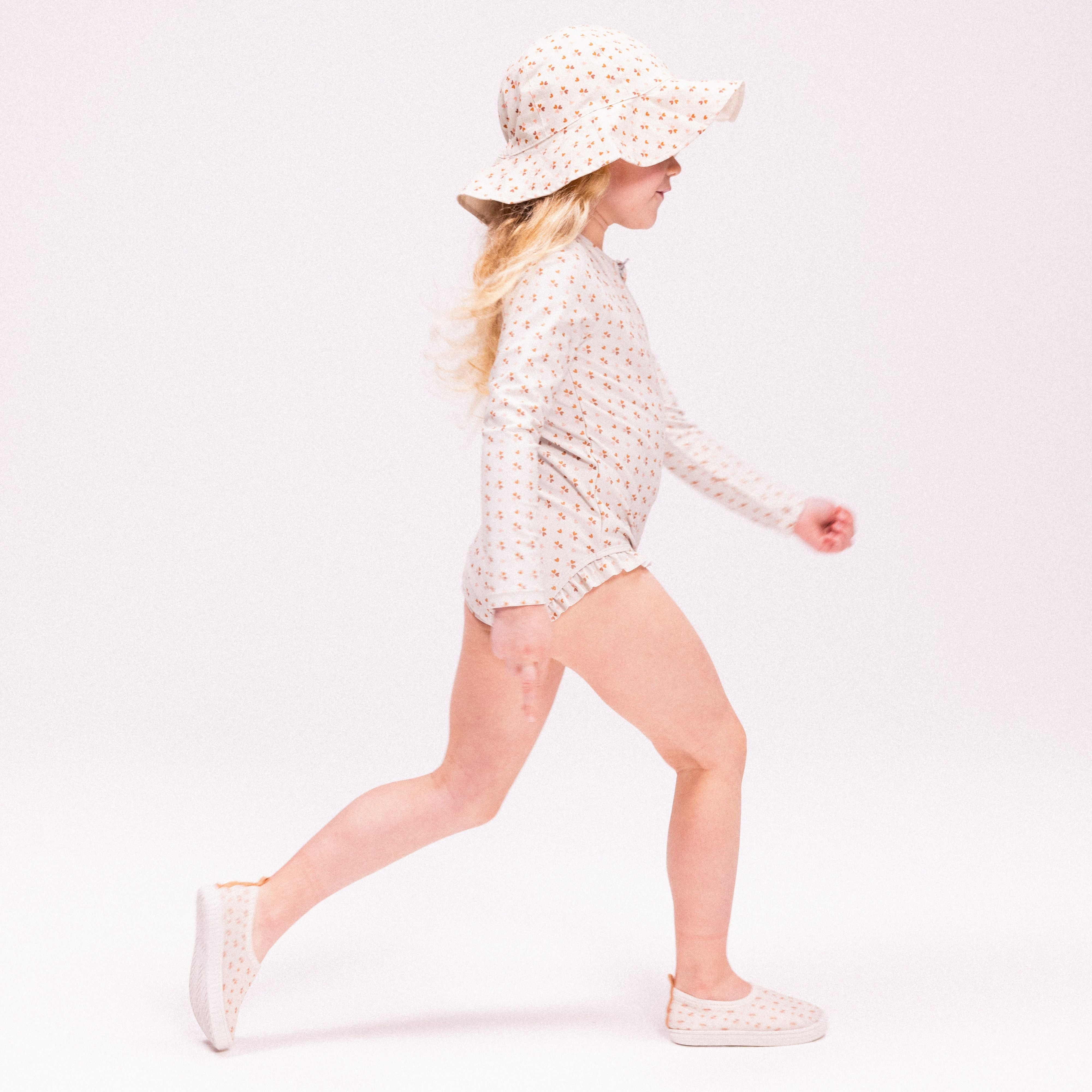A girl wearing an old pink swimsuit with hearts on it with an old pink hat with hearts on a white background