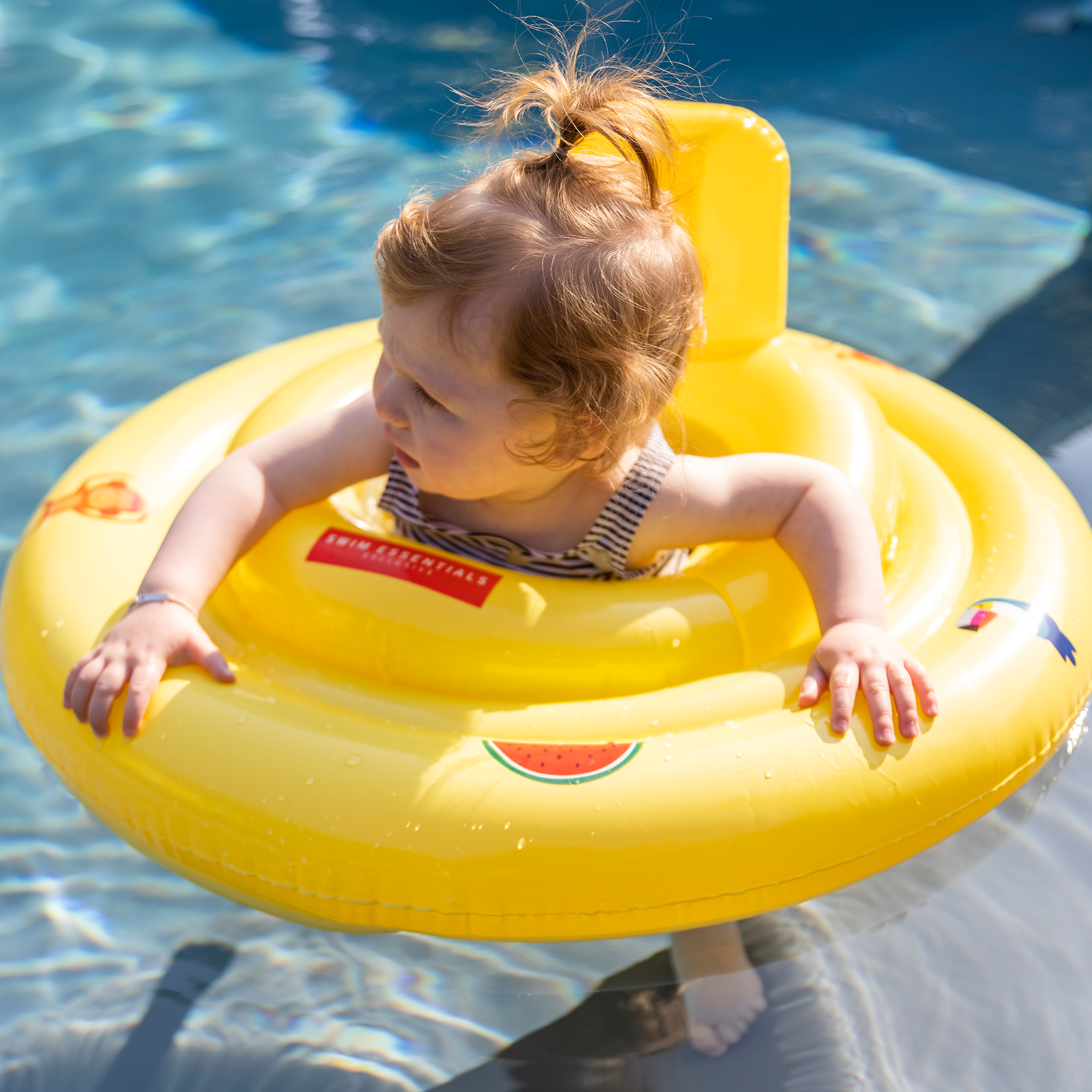 Baby in a yellow Golden Safari Baby Float with cartoons on it, like a watermelon and a monkey in clear blue water
