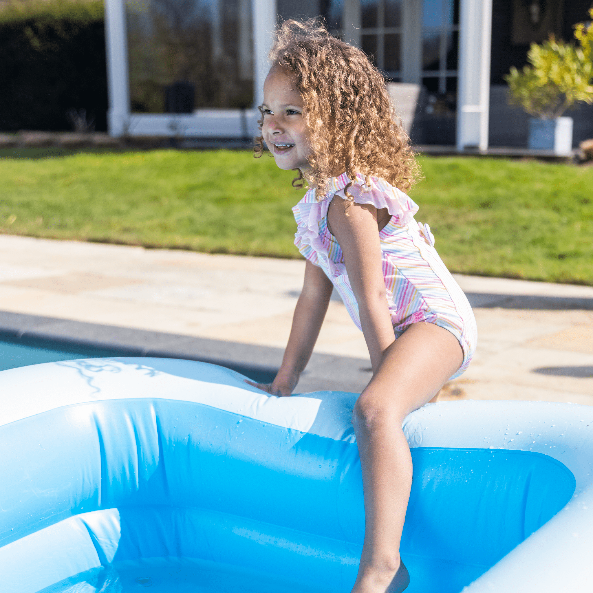 Young girl with curls playing in a blue inflatable pool outdoors with a house in the background.