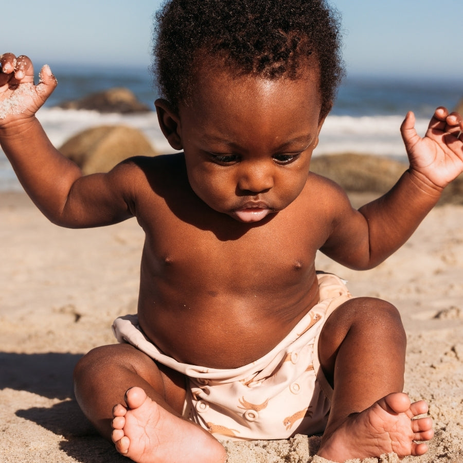 Baby sitting on a sandy beach with ocean in the background wearing a beige swim diaper with animal pattern
