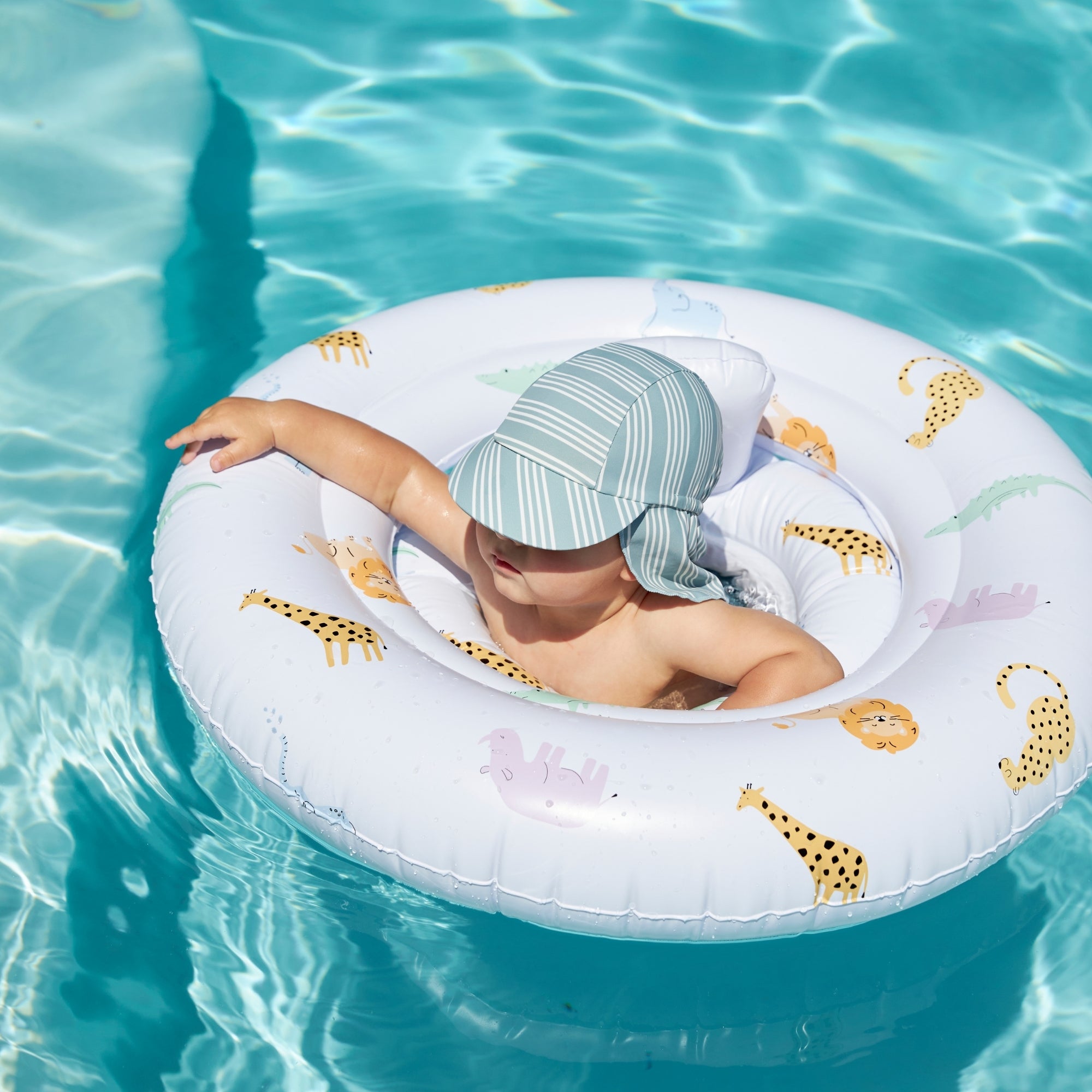A child in a pool sitting in a wildlife-themed baby float. The baby float is white and has an animal print. The child is wearing a blue sun hat with white stripes.