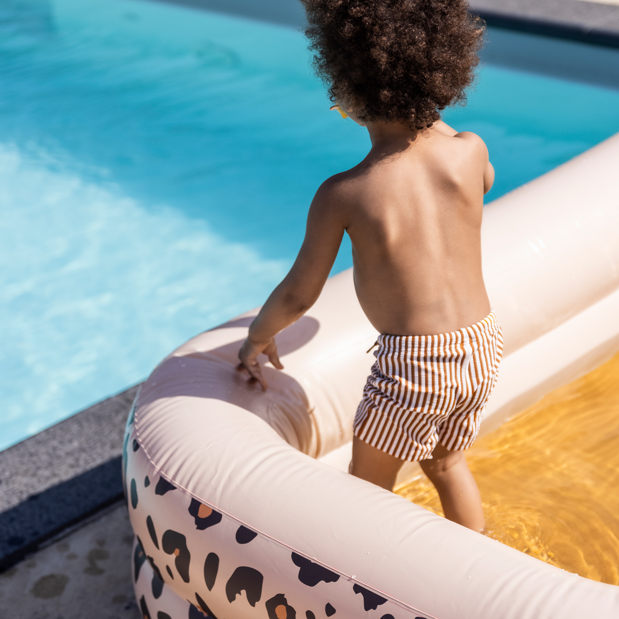 The back of a boy in striped swimming boxer standing in a inflatable pool with leopard print by a pool.