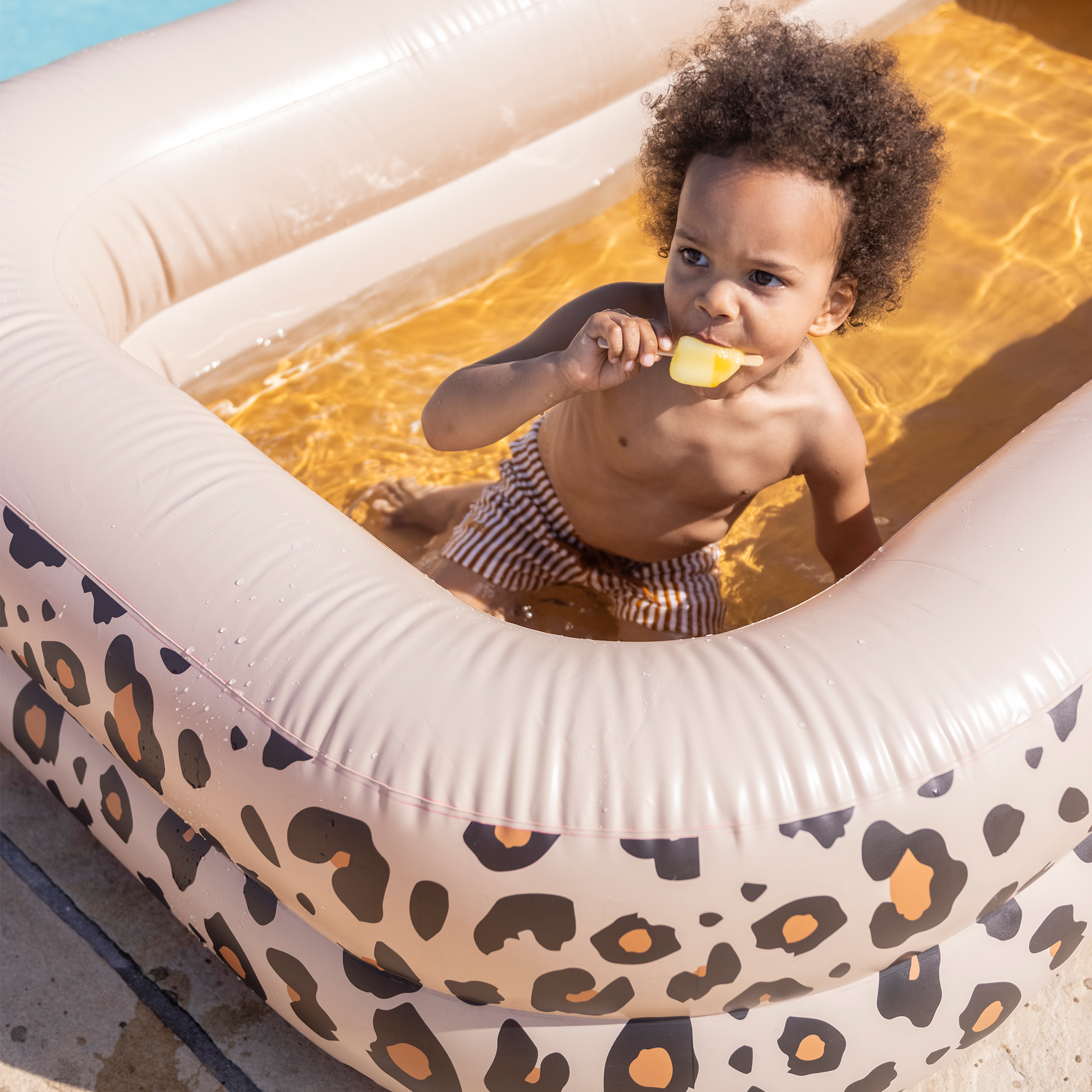 Boy playing in an inflatable pool with a leopard print, holding an ice cream.