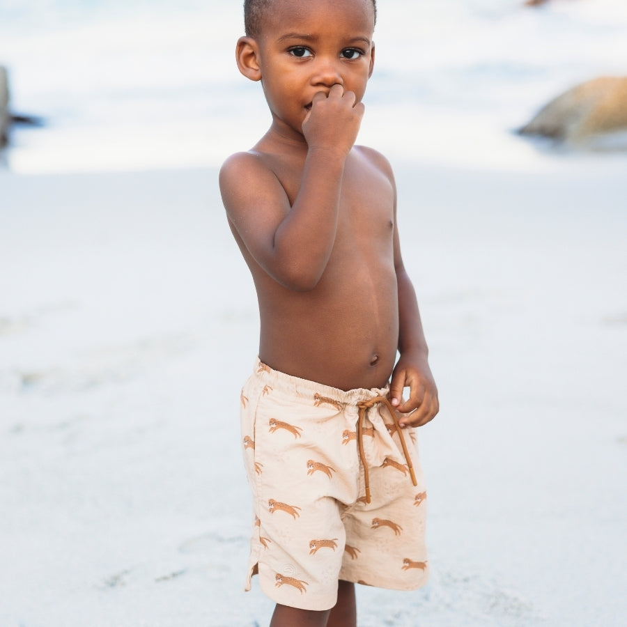Young child wearing beige swim shorts with a pattern, standing on a sandy beach.