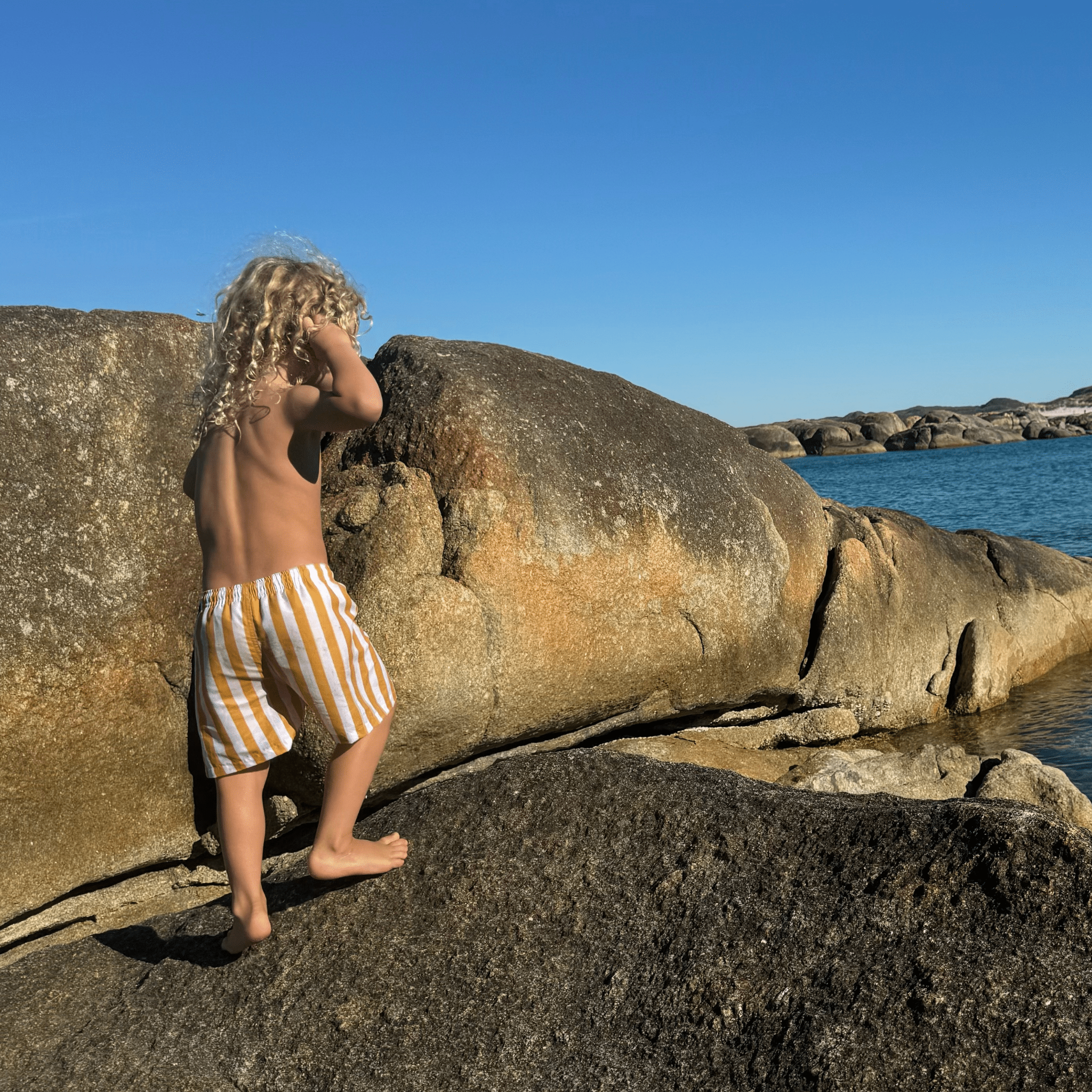 Child in striped swim shorts standing on a rock by the ocean