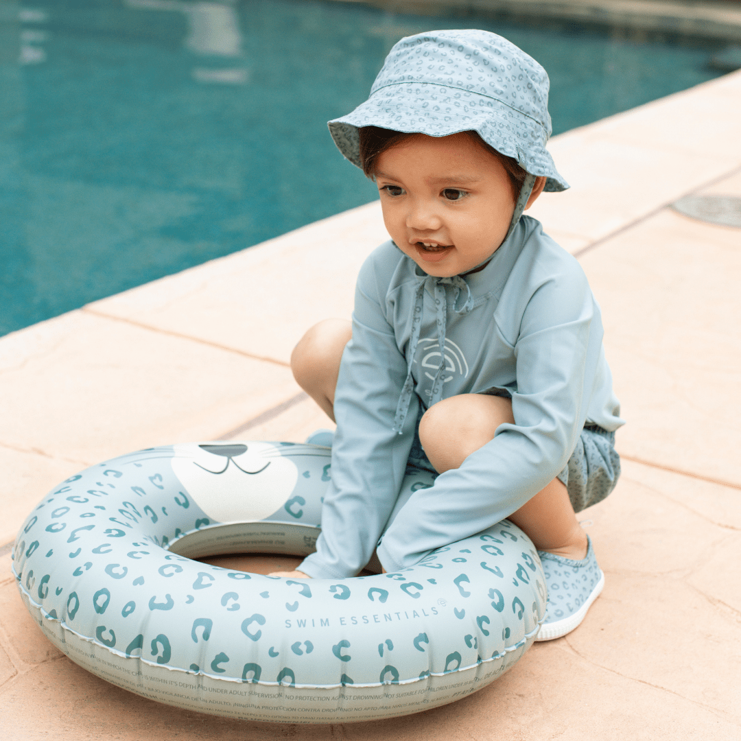 Child wearing a light green outfit and hat, sitting next to an inflatable pool ring by a pool.