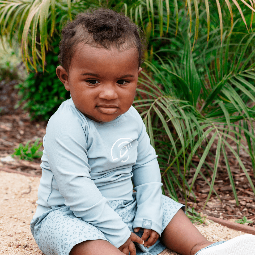 Child wearing a light green long-sleeve shirt and shorts sitting outdoors with plants in the background