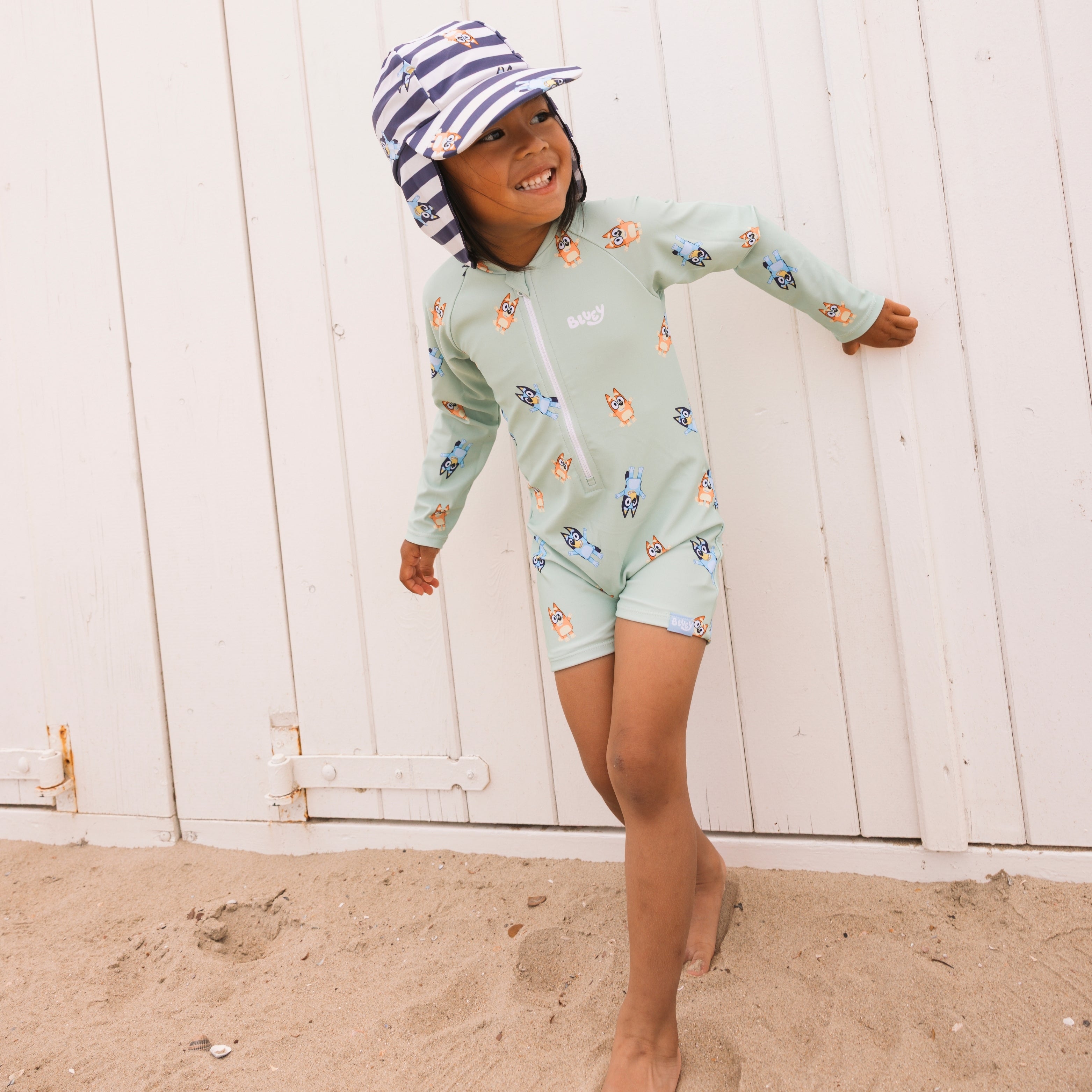 Child wearing a light green swimsuit with cartoon characters and a striped hat on a sandy beach.
