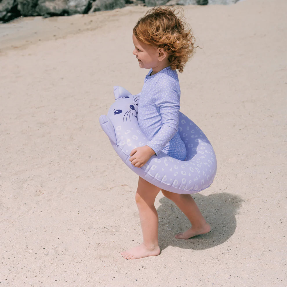 Child photographed from the side wearing a purple leopard swimsuit met long sleeves, a zip and ruffles. Holding a purple leopard inflatable swim ring on a sandy beach.