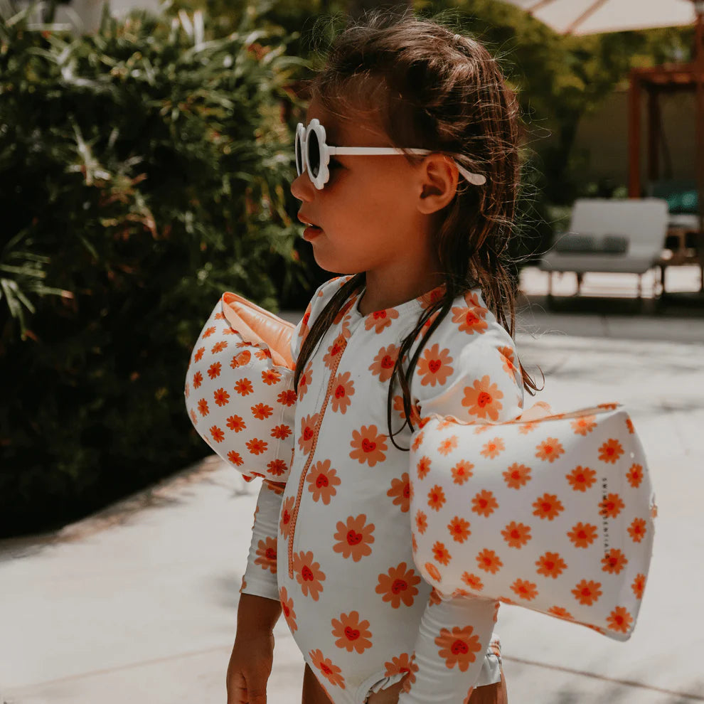 Child wearing a floral swimsuit with long sleeves, a zip and ruffles, wearing with arm bands outdoors.