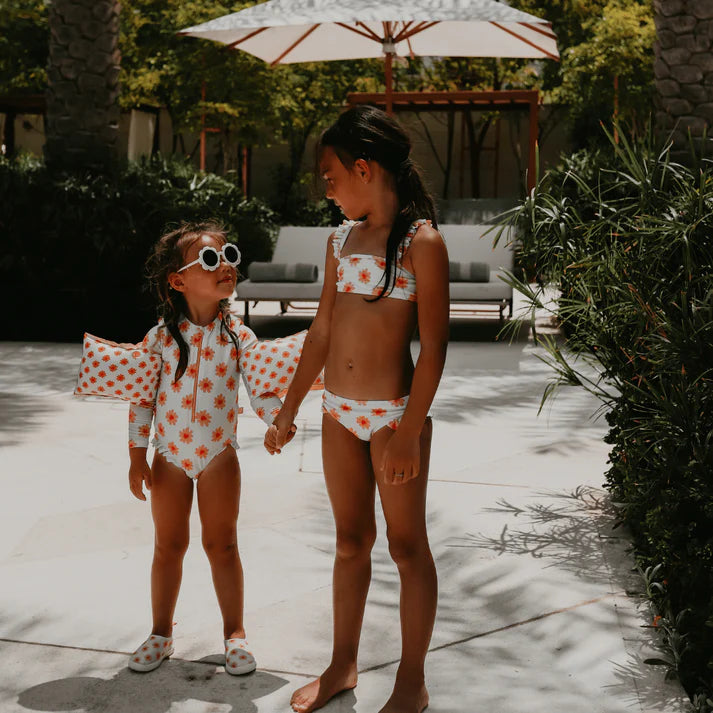 Two young girls in matching floral prints, one in a bikini and one in a swimsuit. The younger girl is also wearing matching water shoes. They are standing outside, holding hands and looking at each other.