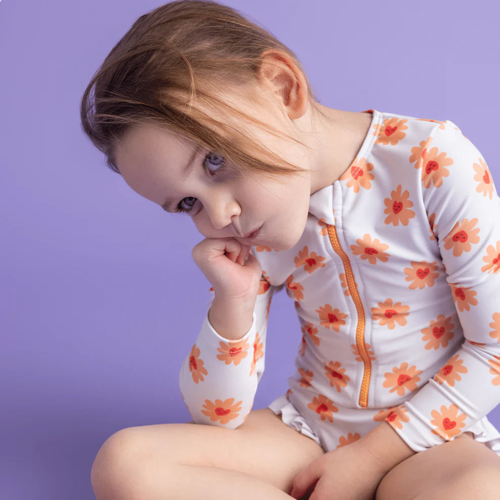 Child sitting on the ground and wearing a floral swimsuit with long sleeves, with a zip and ruffles against a purple background