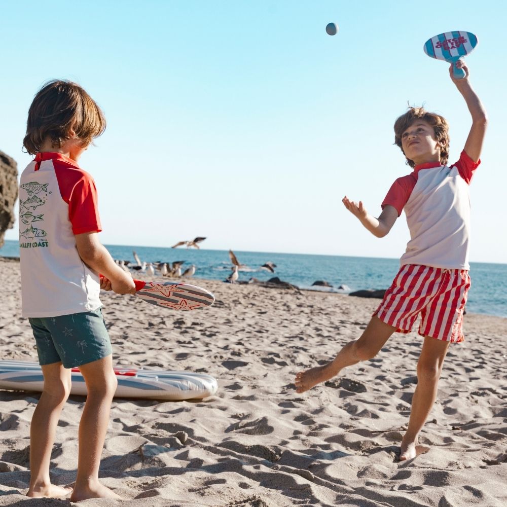 Two boys are playing tennis on a sandy beach, with a clear blue sky and the ocean in the background. One boy is seen from behind; he is wearing a white shirt with a red collar and red short sleeves. Green fish are printed on the back. He is wearing green swim shorts. The other boy is jumping in the air to hit the ball back. He is wearing a white shirt with a red collar and red sleeves. He is also wearing red-and-white striped swim shorts.