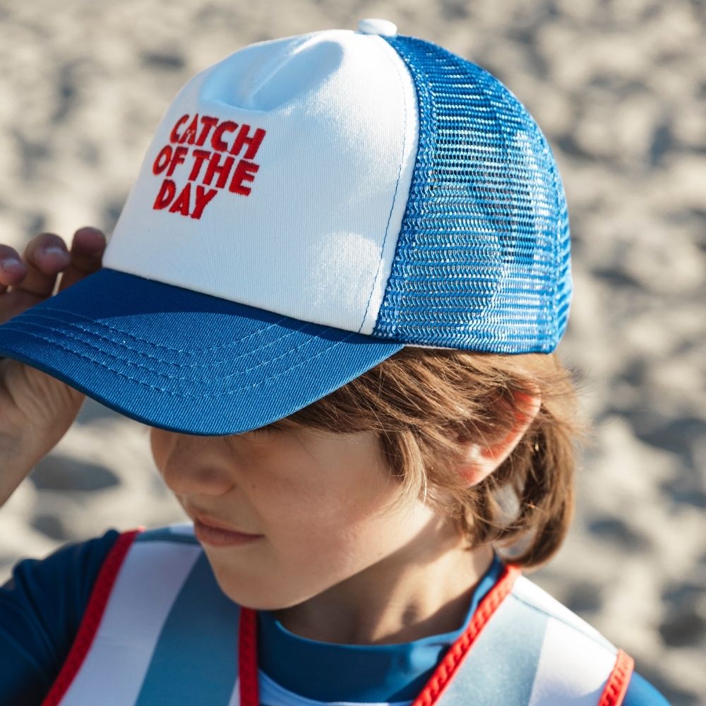 A boy wearing a blue and white cap with 'Catch of the Day' text, standing against a blurred natural background.