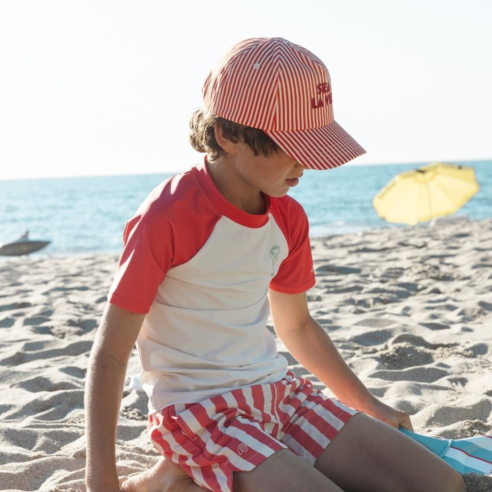 A boy wearing a red-and-white striped cap with the words “Sea la Vie” on it; he is also wearing a white shirt with a red collar and red short sleeves. There is a green lobster in the upper right corner of the shirt. The boy is also wearing red-and-white striped swim shorts. He is sitting on a sandy beach with the ocean in the background.