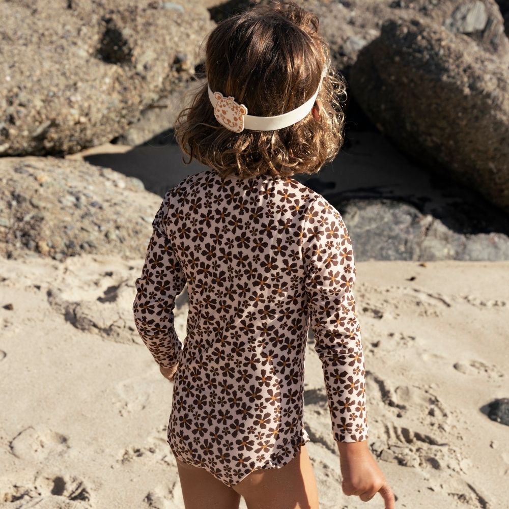 A child is wearing a beige swimsuit with a brown floral print. She is also wearing swimming goggles. She is standing on the beach with a rocky background.