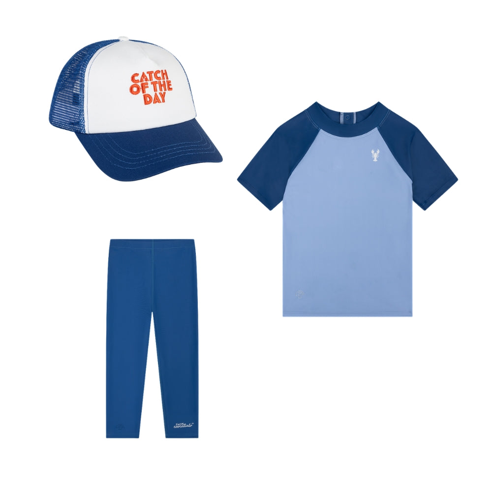 A set comprising a cap, a swim shirt and leggings. The cap is white with blue panels. On the front, on the white panel, the text ‘catch of the day’ is printed in red. The shirt is light blue with dark blue sleeves. The shirt also features a small lobster logo in the top right-hand corner. The leggings are dark blue and feature the Swim Essentials logo in small letters on the front of the right leg at the bottom. The products are shown against a white background.