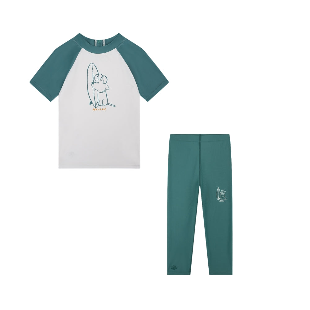 A two-piece children’s outfit comprising a shirt and leggings. The shirt is white with green sleeves. The white section features a dog with a surfboard, along with the text ‘sea la vie’. The leggings are green and, in the middle of the right leg, there is a surfboard with a wave and a palm tree on a white background.