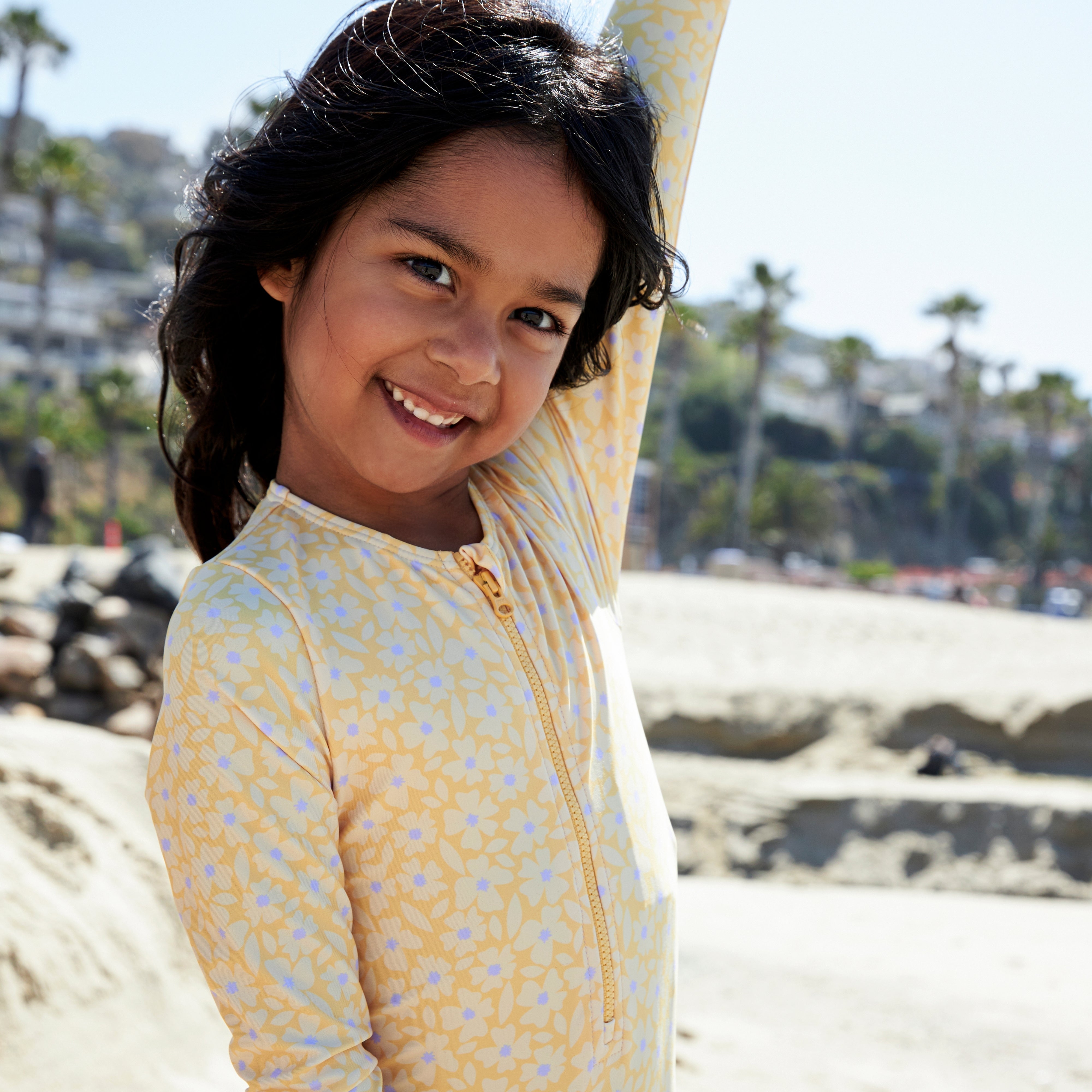 Young girl in a yellow patterned outfit standing on a beach with palm trees in the background.