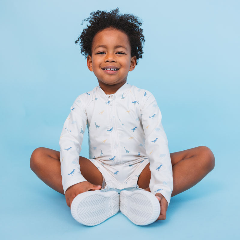 A child is sitting on the floor wearing a white swimsuit with a dinosaur print. He is also wearing water shoes. Against a light blue background. 