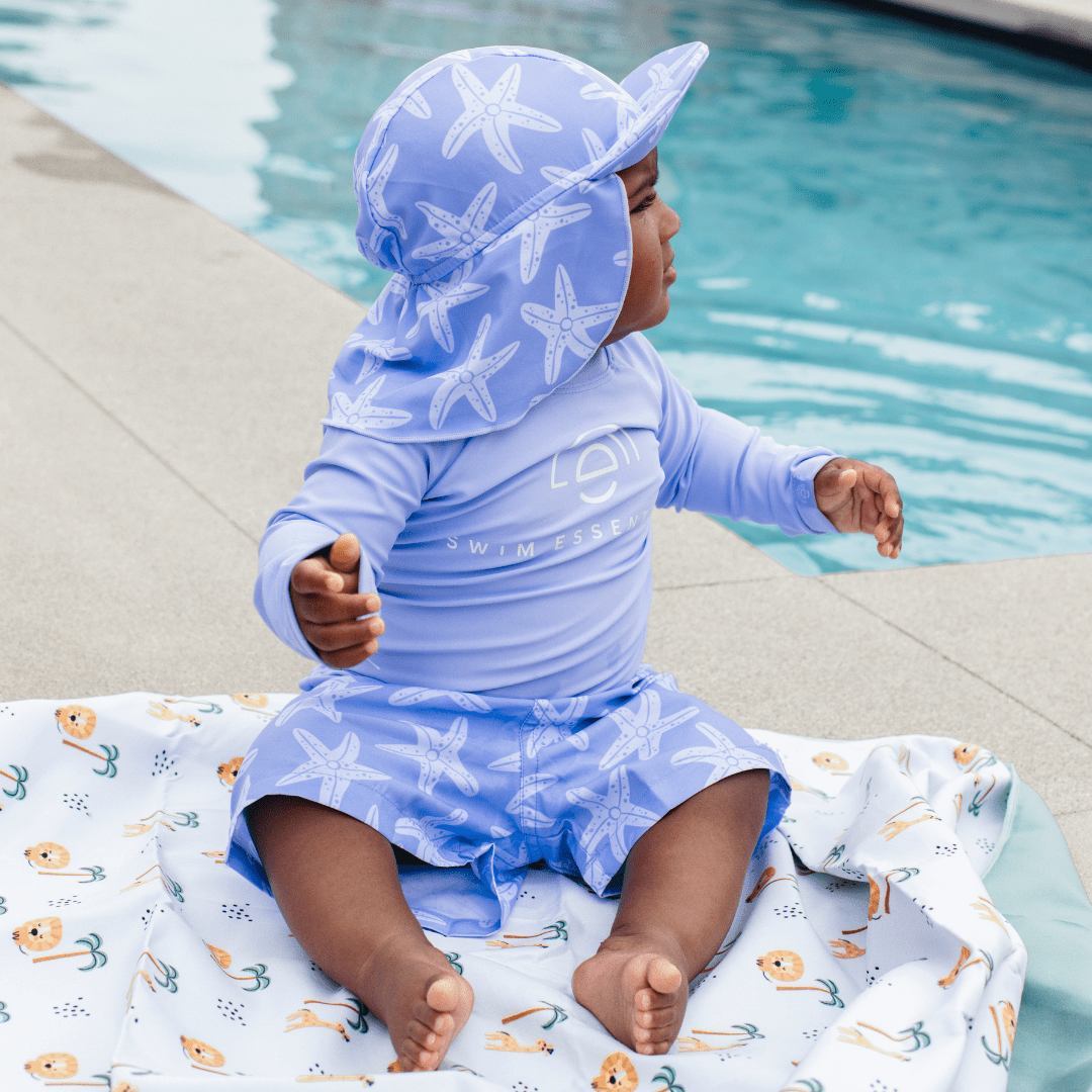 Child in a starfish-themed swimsuit and hat sitting next to a pool and in a towel