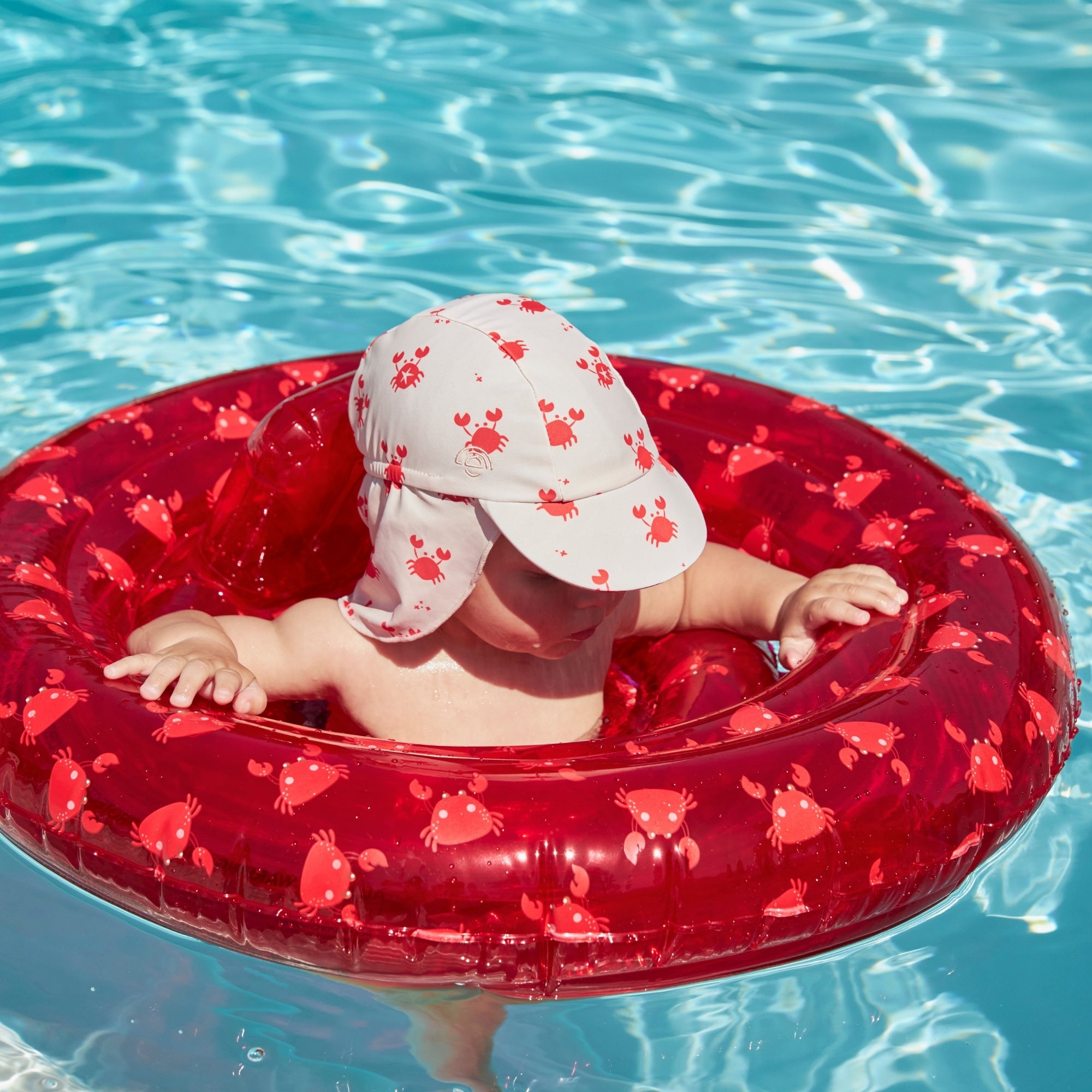 Small child who is wearing a cap with crabs on it, looking down in a transparant red baby float with crabs on it in clear blue water. 