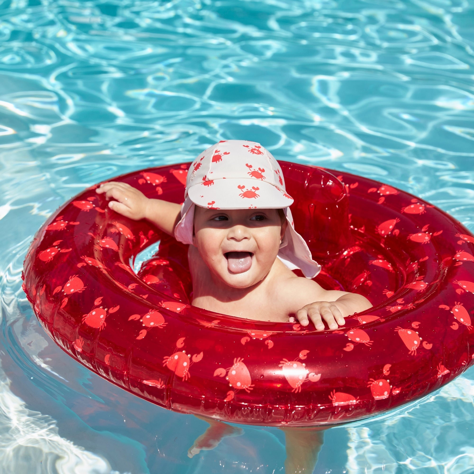 A child in a pool sitting in a transparent red baby float with a crab print. The child is wearing a beige cap with sun protection at the back of the neck. The cap has a crab print. 