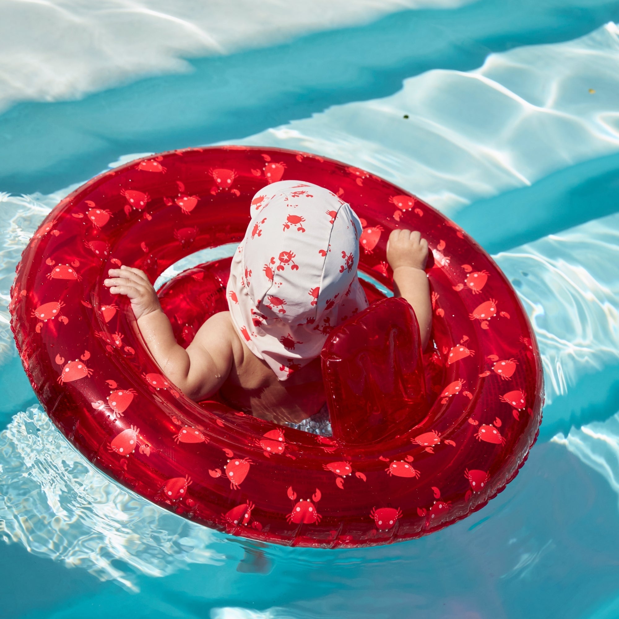 Child in a red baby float inflatable floating in clear blue water with the crabby sun cap
