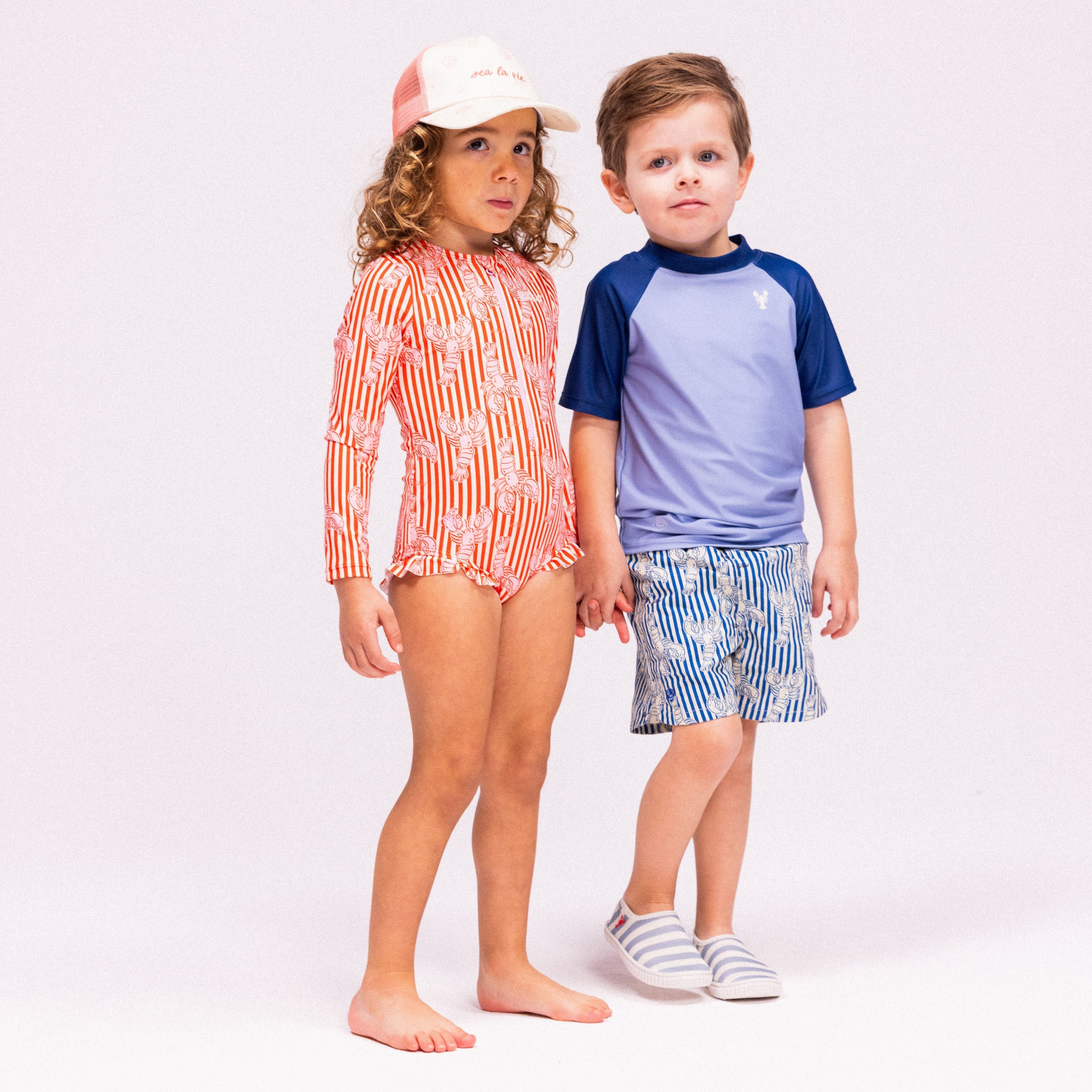 Two children are standing hand in hand. The girl is wearing a white cap with the text "Sea la Vie" and a swimsuit with red and white stripes and a lobster print. The boy is wearing a light blue swim shirt with a dark blue collar and dark blue short sleeves. His swimming trunks are white with blue stripes and a lobster print. He is also wearing white and blue striped water shoes. The background is gray.