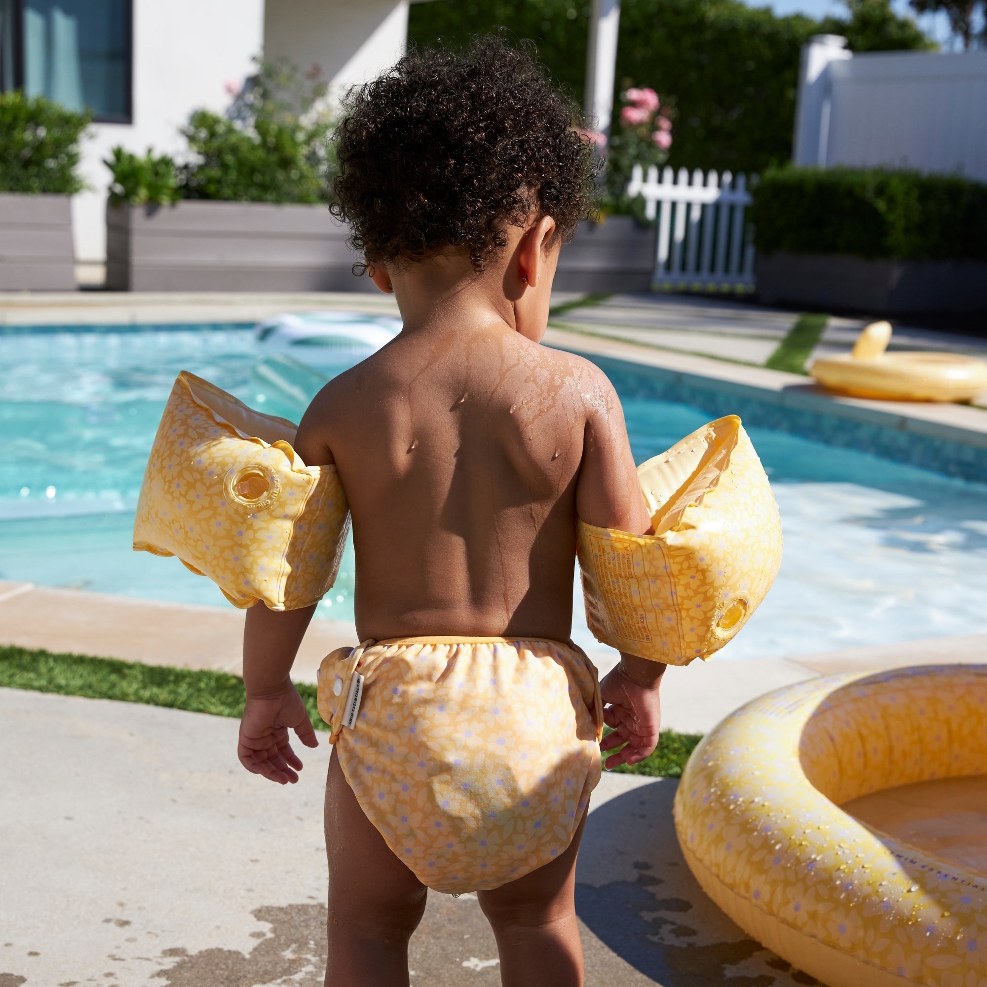 Child with yellow arm floats and swim diaper standing by a pool