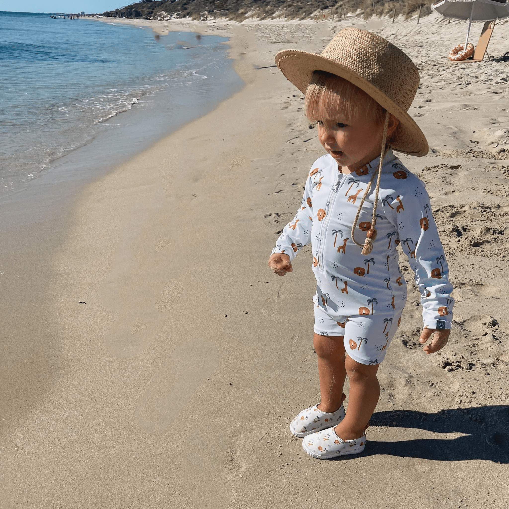 Child in a patterned swimsuit, water shoes & sun hat standing on a sandy beach.