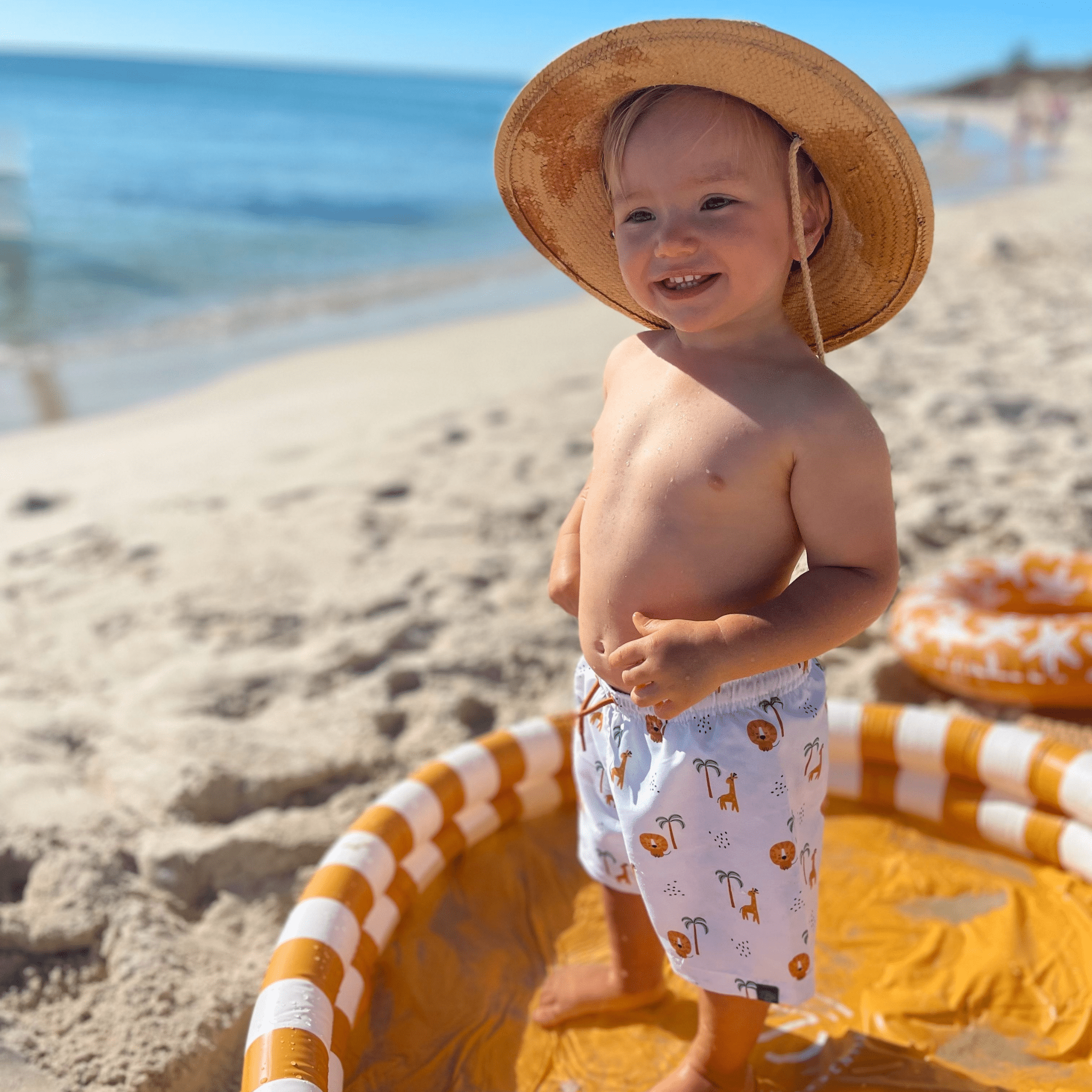 Child on a beach wearing a straw hat and swim shorts with a pool float in the foreground.