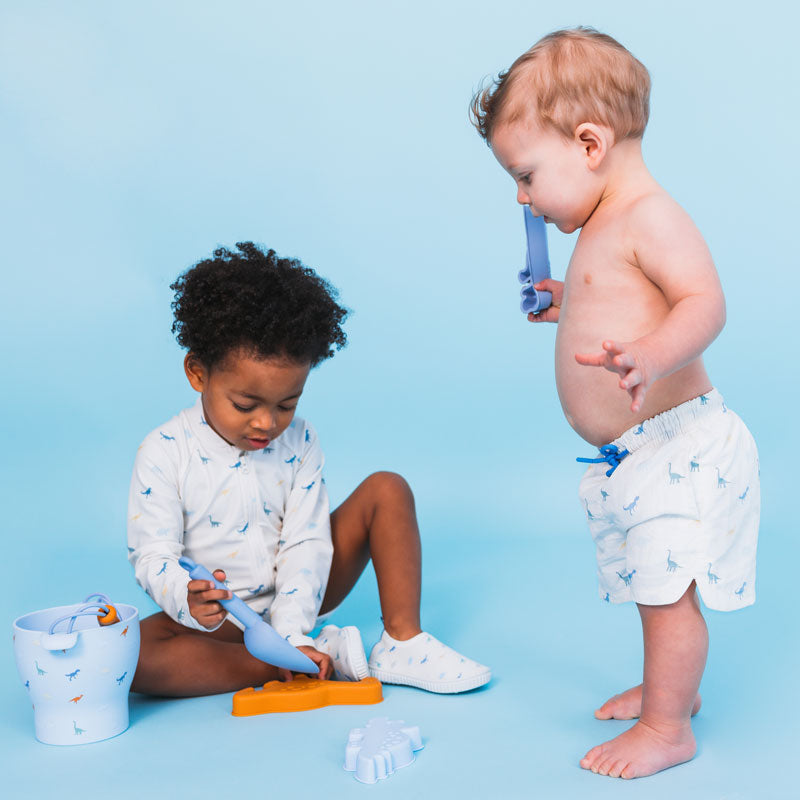 Two children playing with toys on a blue background wearing swimwear with a dino print