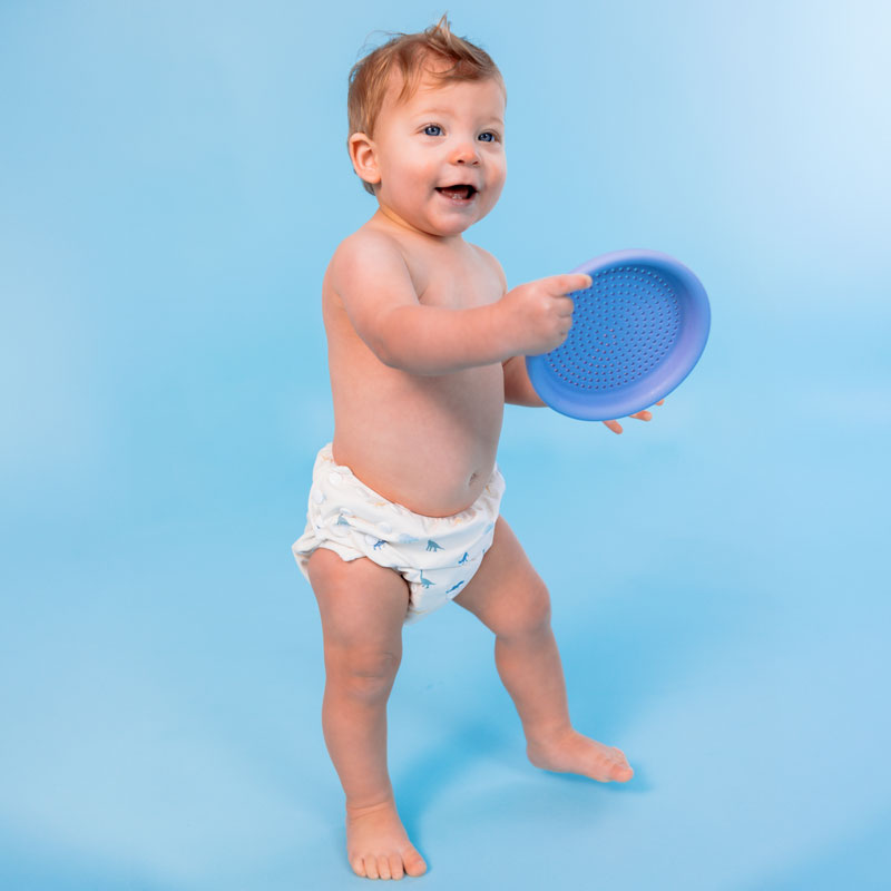 Baby holding a blue plastic toy against a light blue background wearing a swim diaper with dinosaur pattern