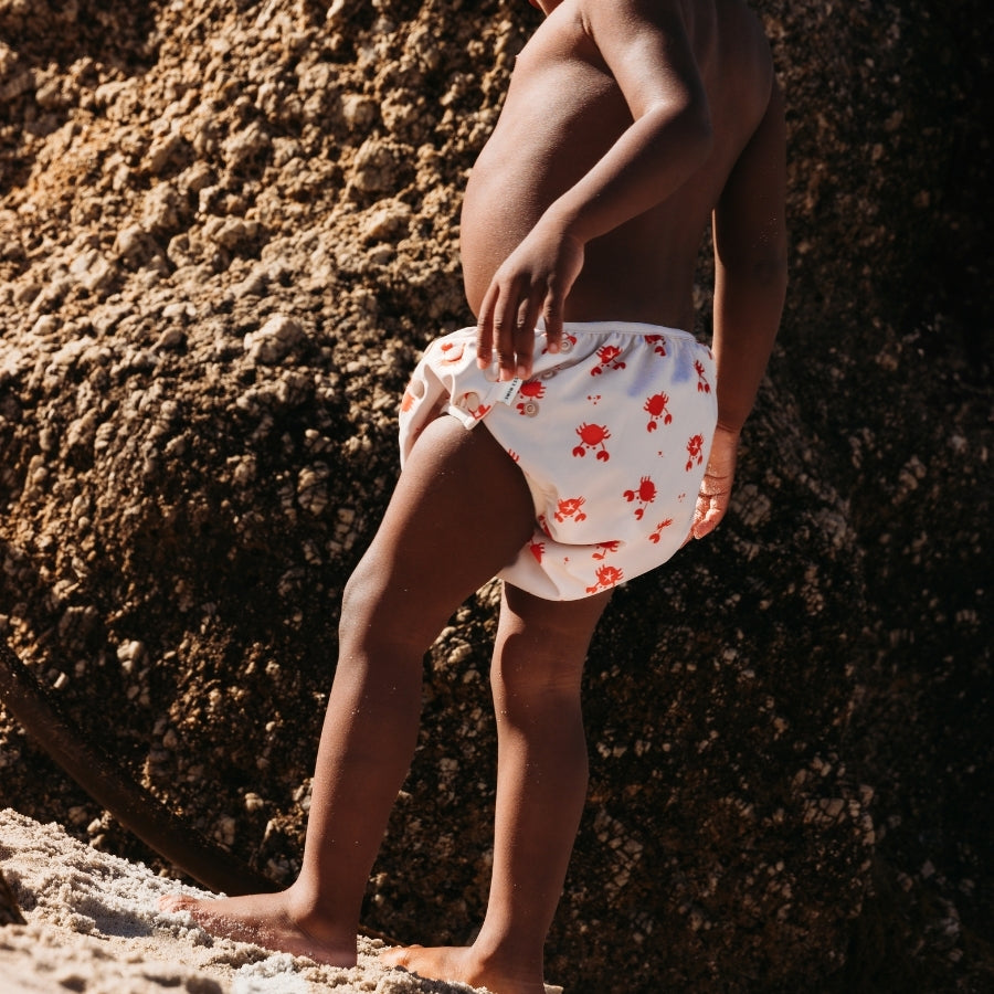 Baby wearing beige swim diaper with red crab pattern standing on sand with rocks in the background
