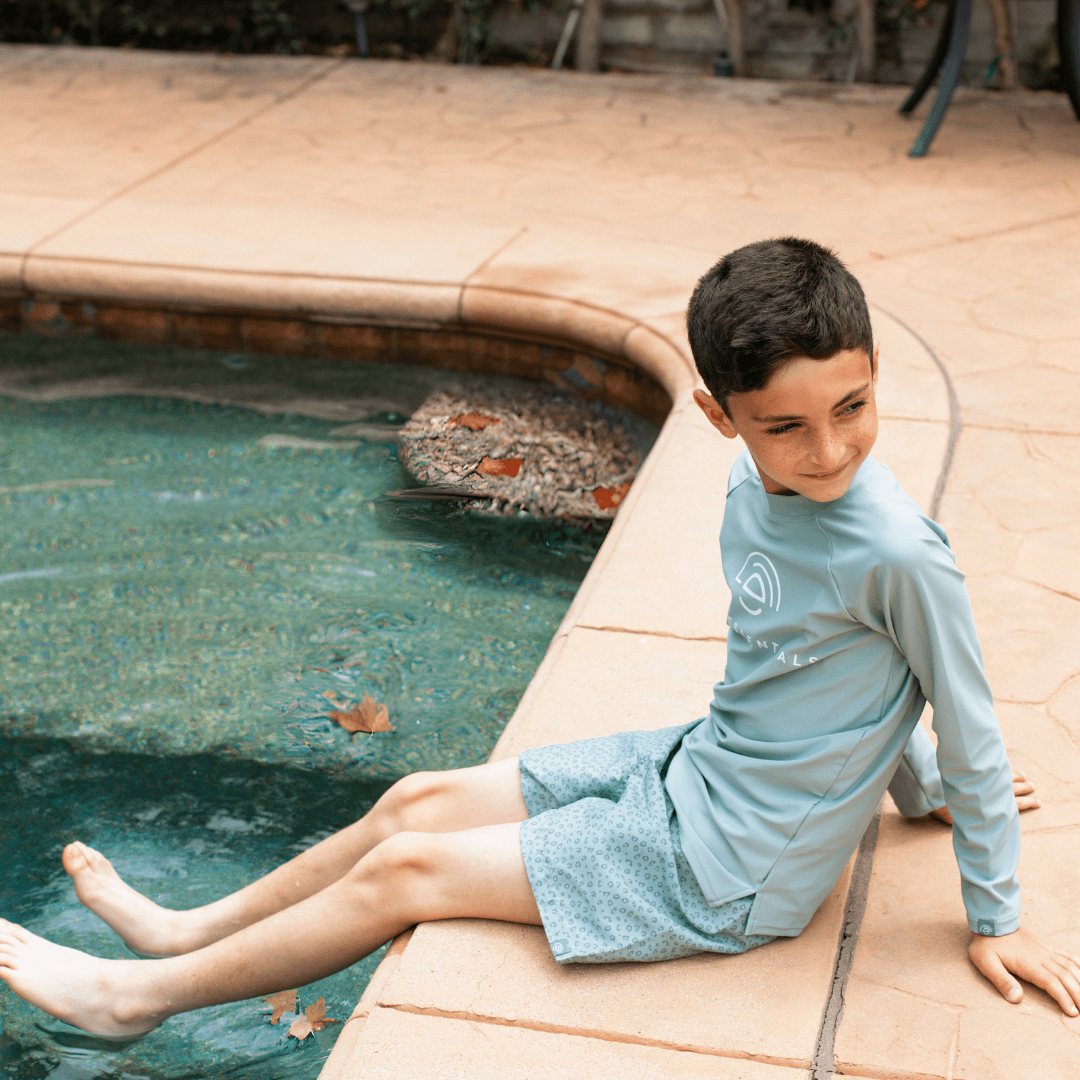 Child sitting by a pool wearing a light green long sleeve swim t-shirt with a white 'Swim Essentials' logo.