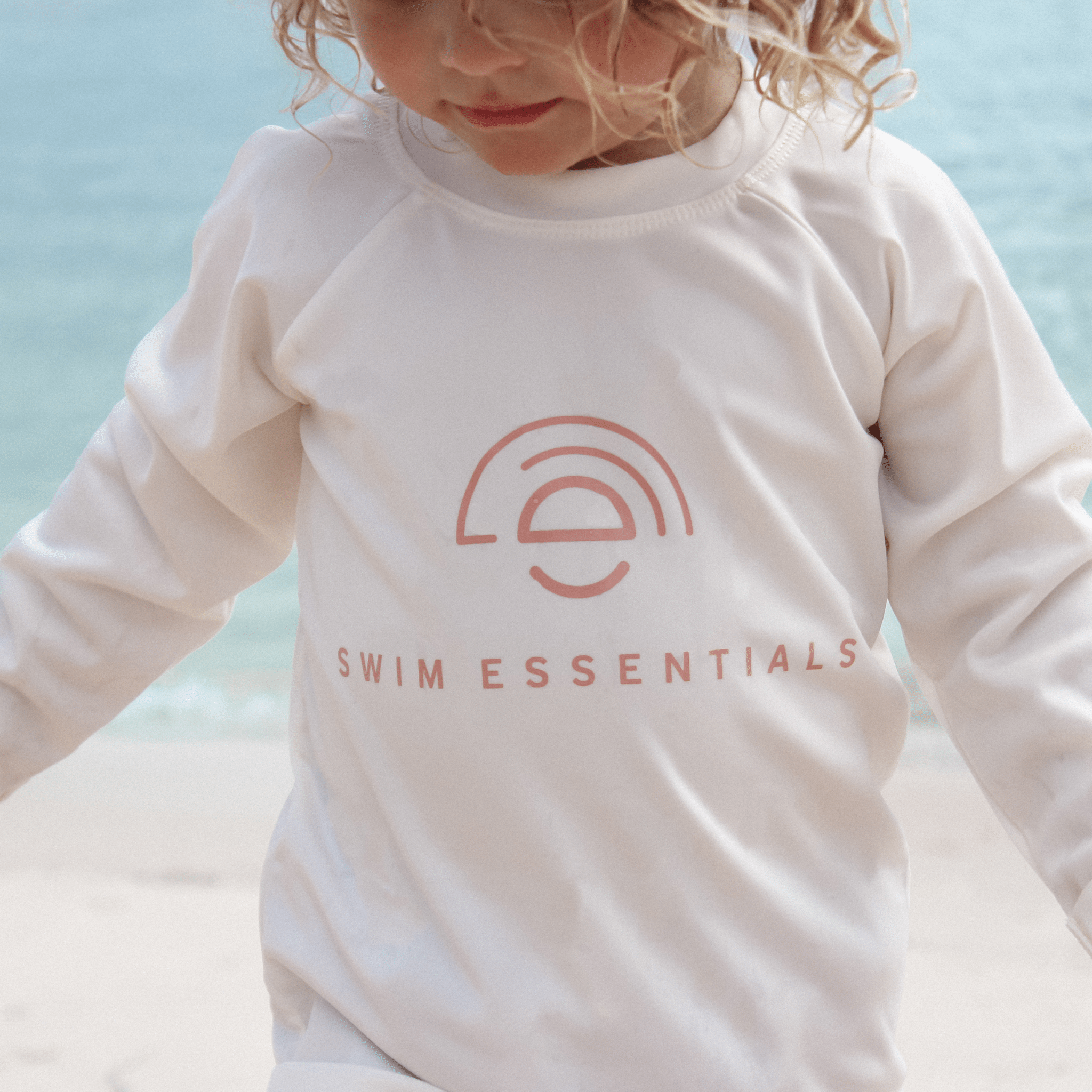 Child wearing a white long-sleeve shirt with a pink'Swim Essentials' logo against a light blue background