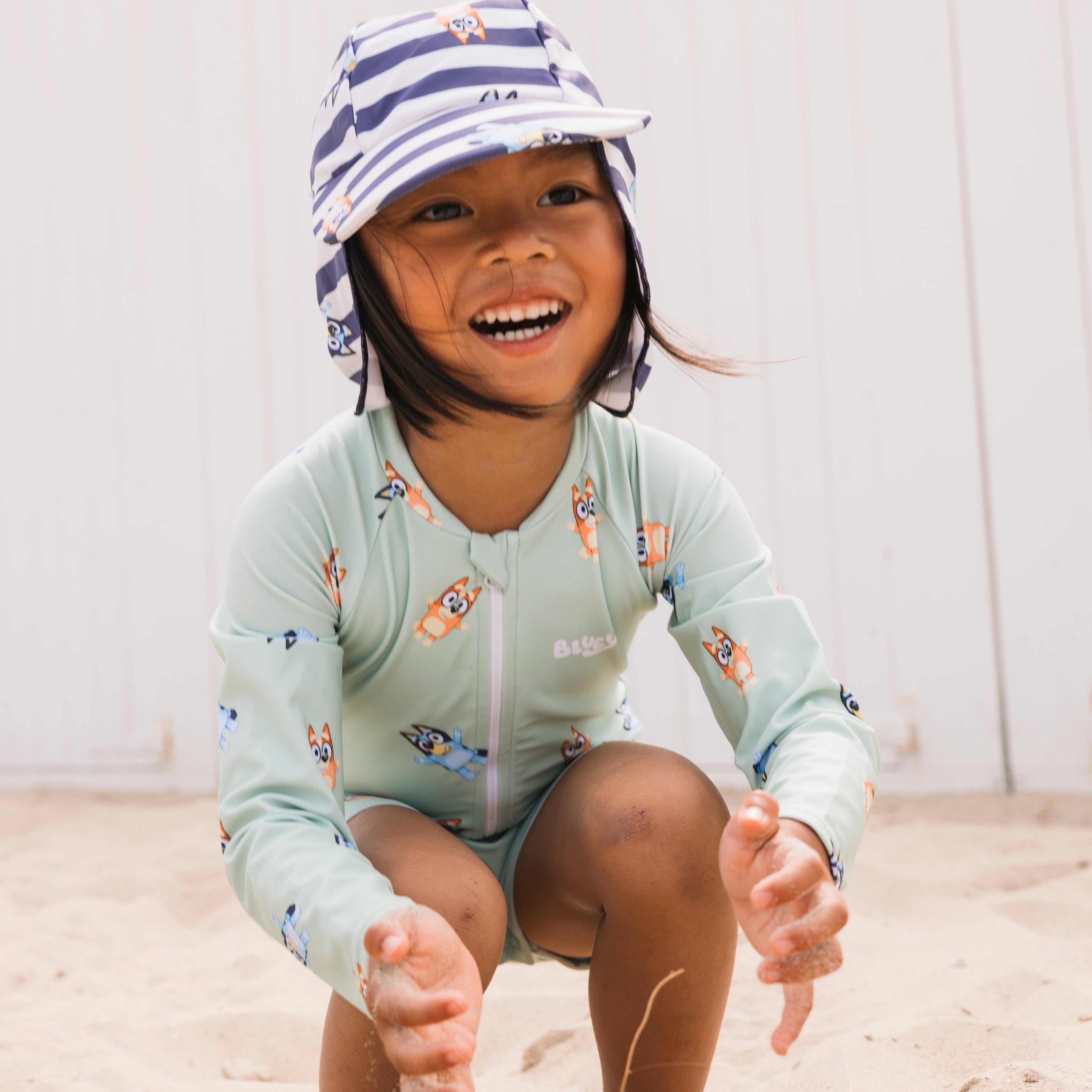 Child wearing a light blue long-sleeve shirt and striped hat, sitting on a sandy surface.