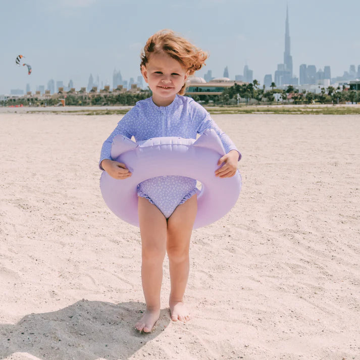 Child wearing a purple leopard swimsuit met long sleeves, a zip and ruffles. Holding a purple inflatable swim ring on a sandy beach with a city skyline in the background