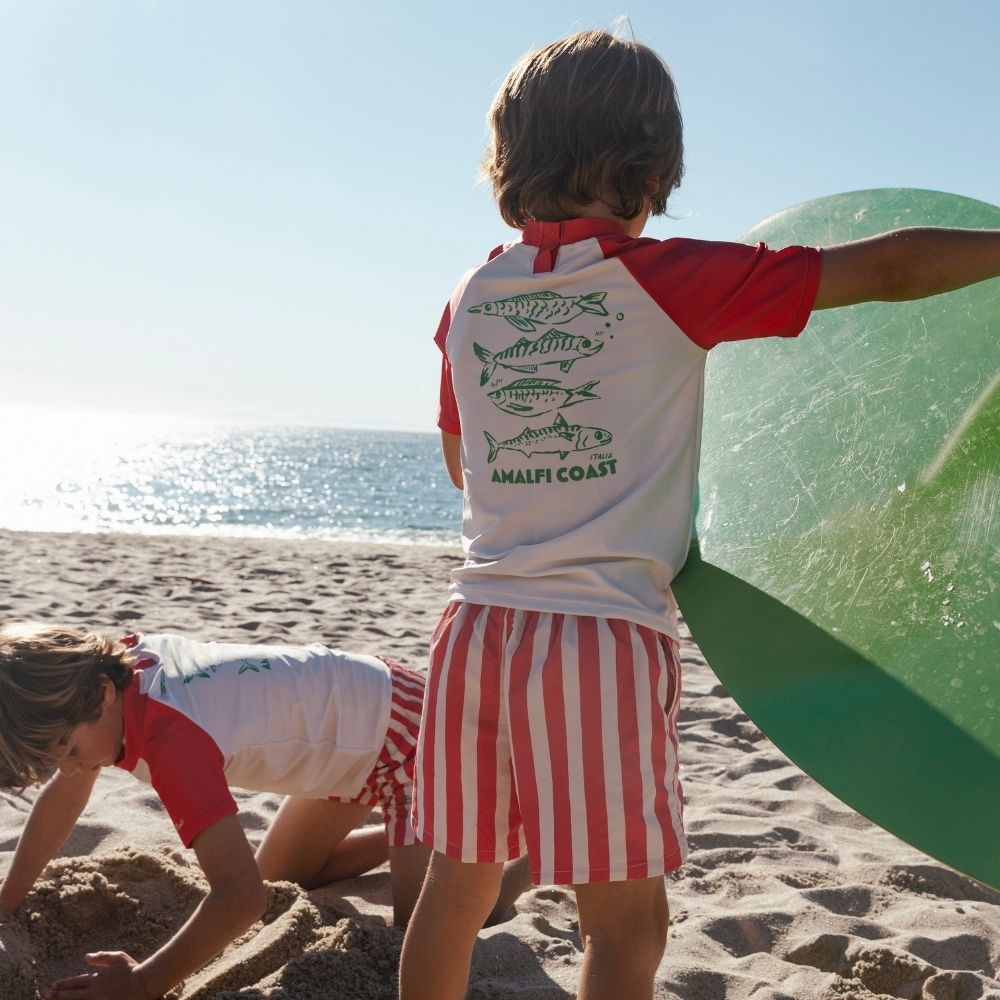 Two boys are playing on a sandy beach under a clear blue sky. Both boys are wearing white swim shirts with red collars and pink short sleeves, featuring green fish on the back and the text “Amalfi Coast.” They are also both wearing red-and-white striped swim shorts. One boy is standing in the sand and the other is kneeling. 
