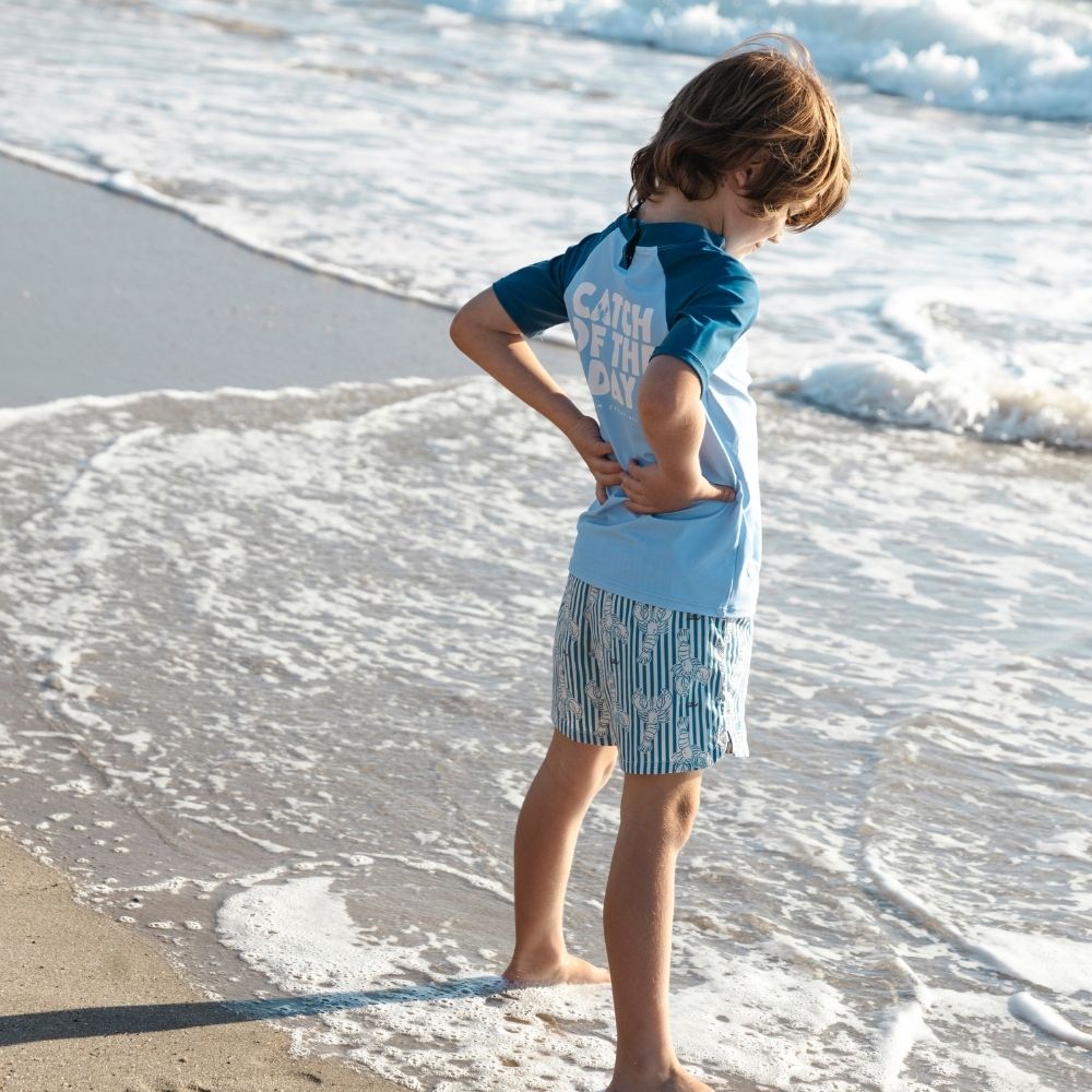 A boy wearing a blue short-sleeved shirt. The back of the shirt reads “Catch of the day.” He is also wearing blue-and-white striped swim shorts with a lobster pattern. He is standing on a beach with waves.