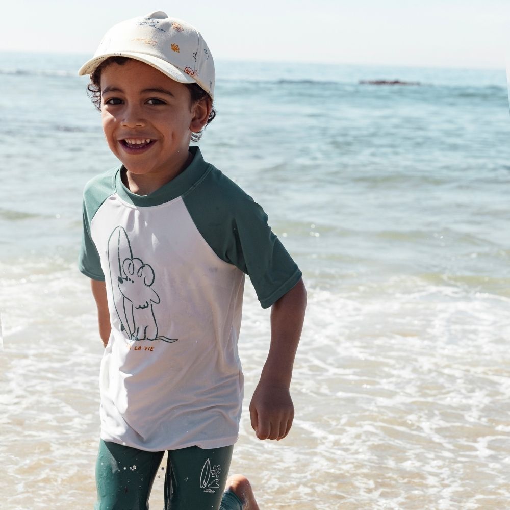 A boy is wearing a cap with a surfer print and a white swim shirt with an image of a dog and a surfboard, with green short sleeves and a green collar. He is also wearing green leggings with a white logo of a surfboard and a wave. He is standing on the beach.


