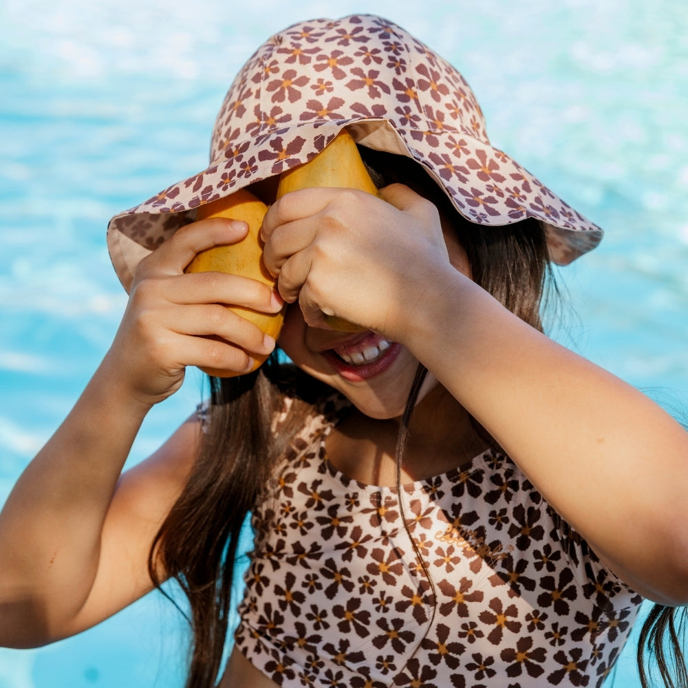 A girl wearing a beige sun hat with a floral print and a beige bikini with a floral print. She is covering her face with her hands. A swimming pool can be seen in the background.